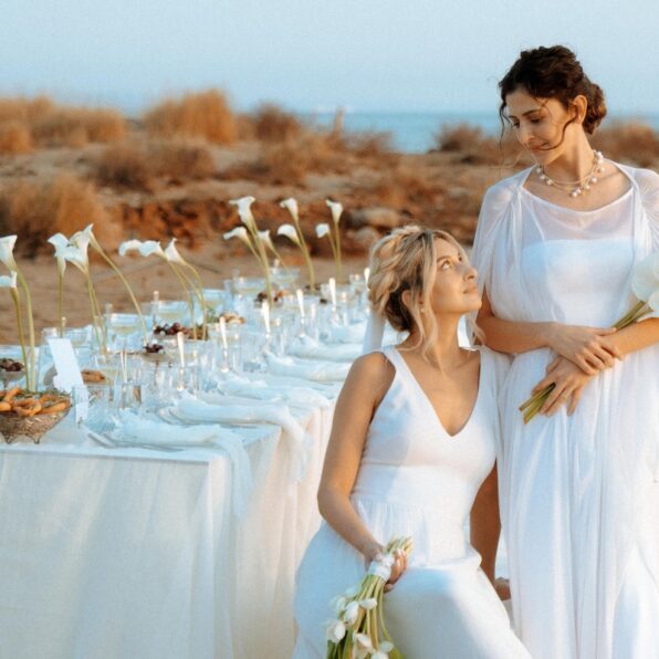 Two women in white dresses pose beside an outdoor table set with white flowers and tableware on a beach, with dry grass and the sea in the background.