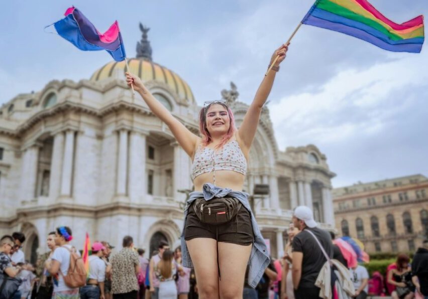 A person stands in front of a historic building, holding up two pride flags, surrounded by a crowd during a public event.