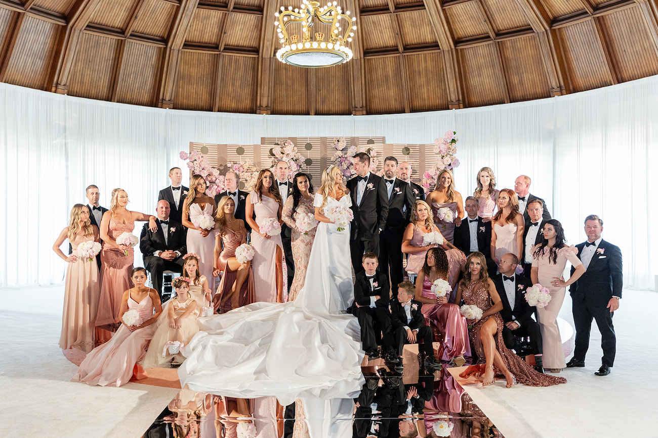 A large wedding party poses indoors, with bridesmaids in pink, groomsmen in black suits, and a bride and groom standing at the center under a chandelier.