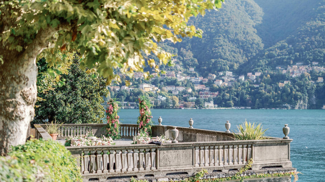 A stone terrace with floral decorations overlooks a lake and distant hillside town, surrounded by greenery under a partly shaded sky.