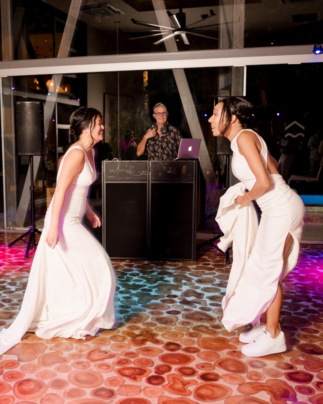 Two women in white dresses dance and laugh on a patterned floor under colorful lights.