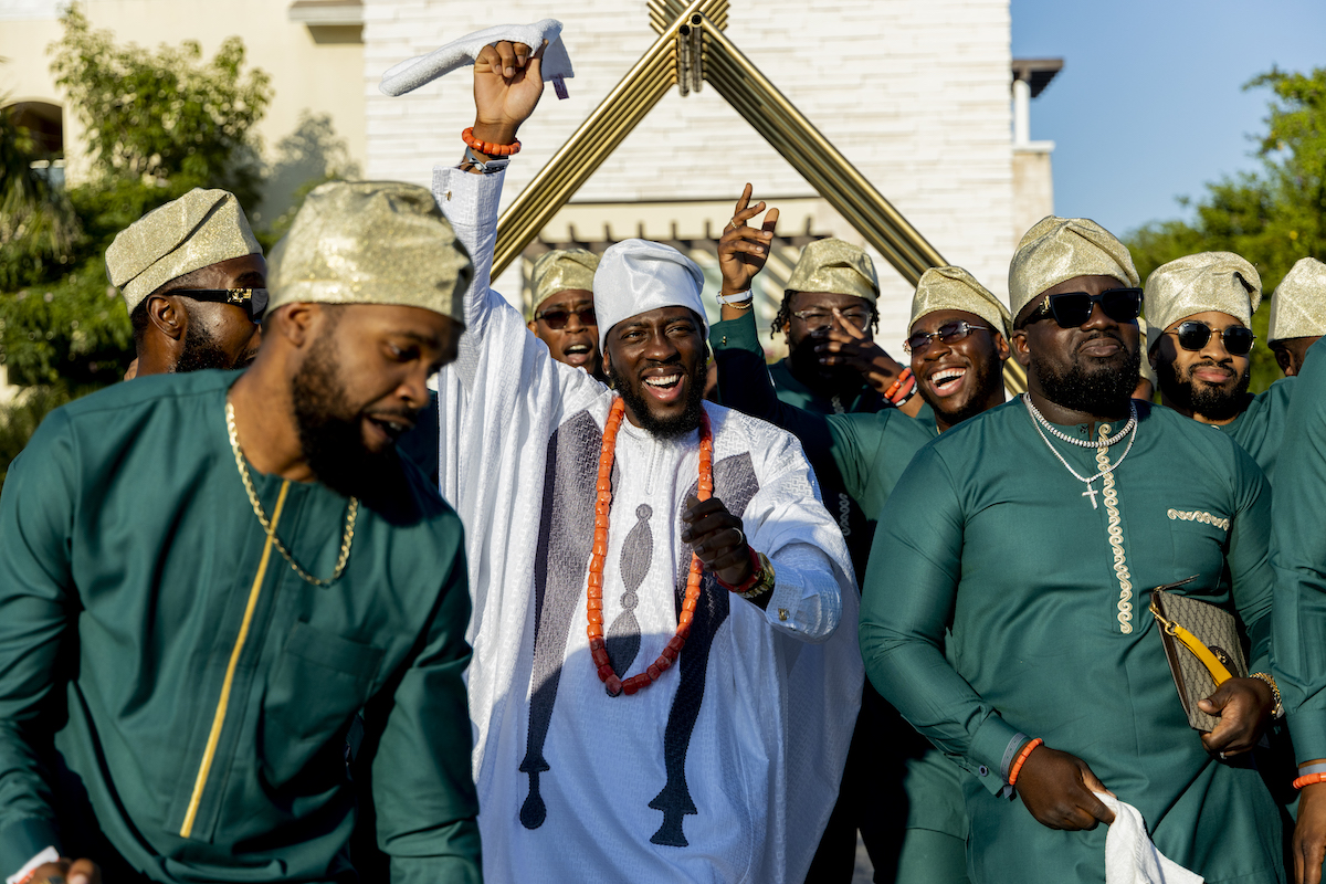 A group of men dressed in traditional attire celebrate outdoors, with one man in white raising his arm and smiling, surrounded by others in green outfits and gold caps.