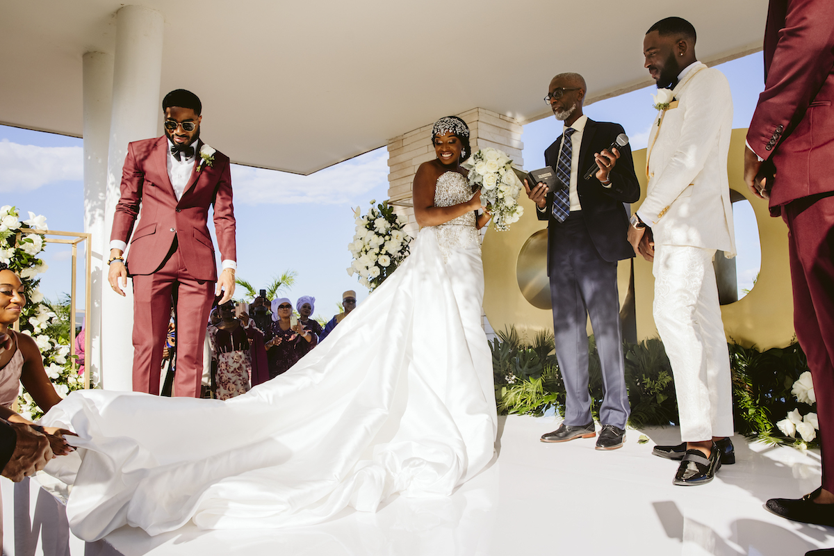 A bride in a white gown stands at the altar with her groom in a white suit, an officiant, and two groomsmen in burgundy suits during a wedding ceremony.
