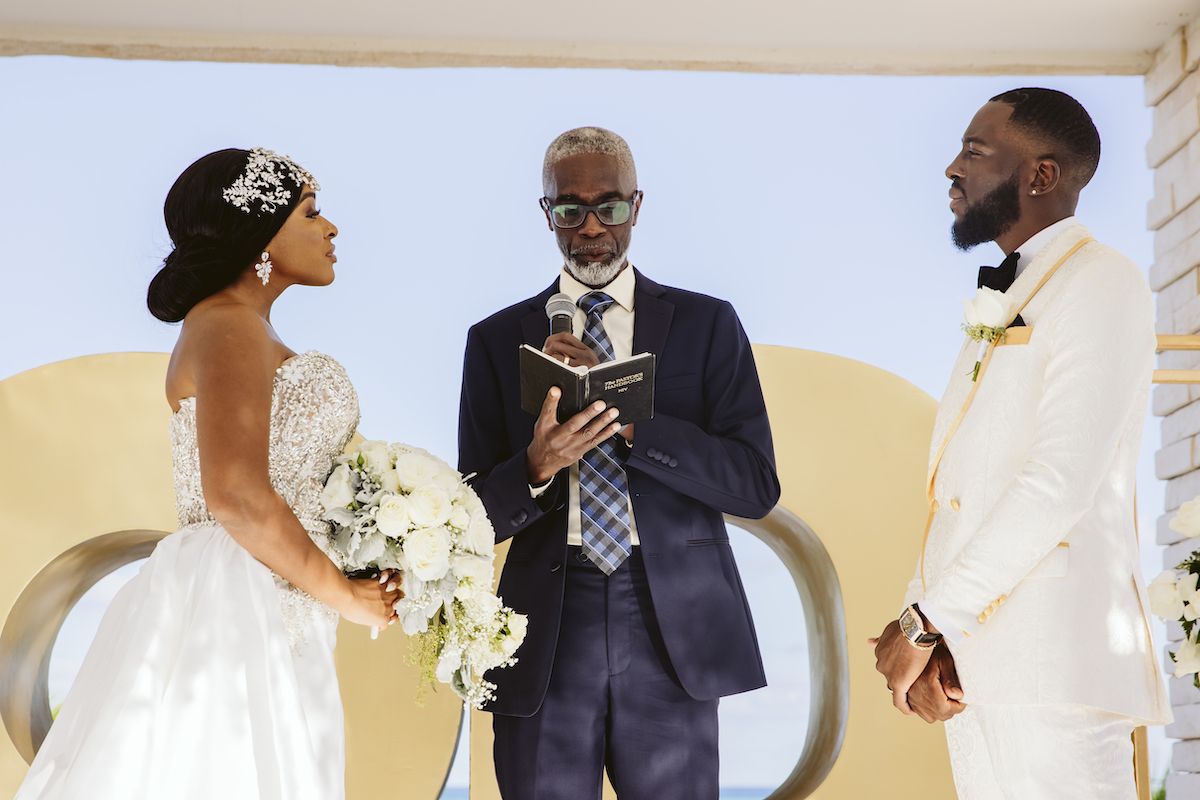 A bride and groom stand facing each other at an outdoor wedding ceremony while an officiant reads from a book.