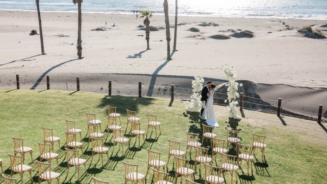 A couple stands under a floral wedding arch on a grassy area near the beach, with empty chairs arranged in rows and palm trees along the shoreline.
