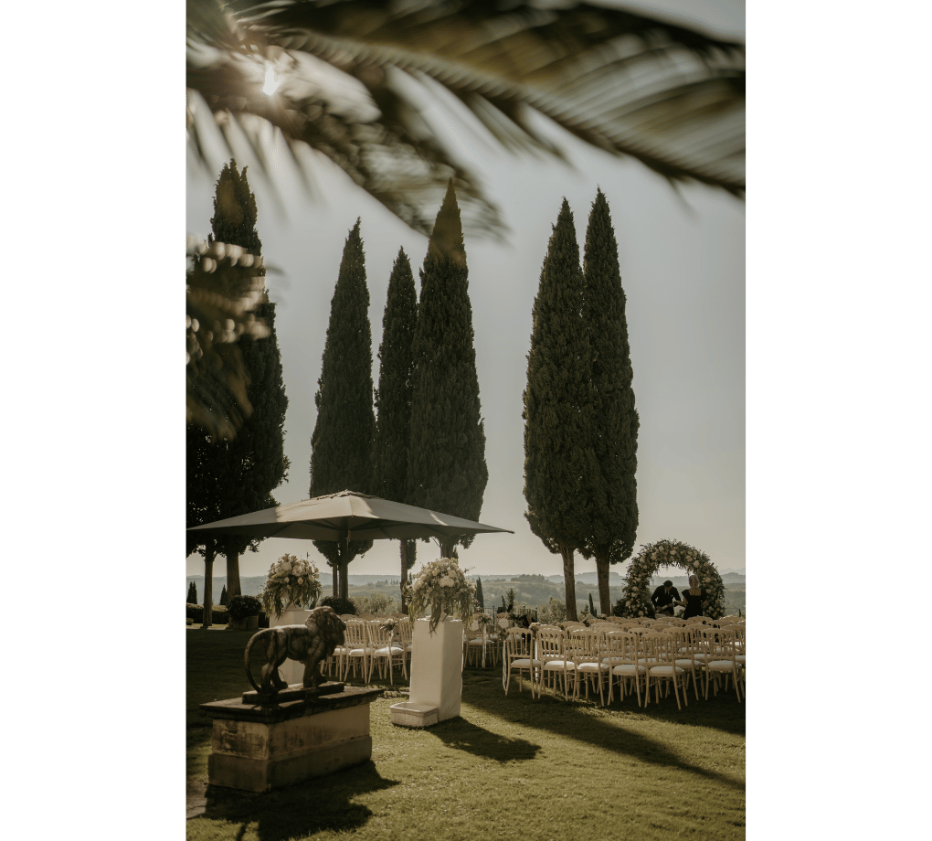 Outdoor event setup with rows of chairs, a floral arch, statue, and tall cypress trees under filtered sunlight. This wedding was planned by Bweddings.