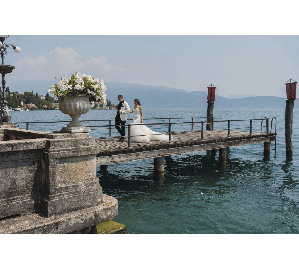 A bride and groom stand together on a wooden pier over a body of water, with stone railings and a large flower arrangement nearby. This wedding was planned by Bweddings.