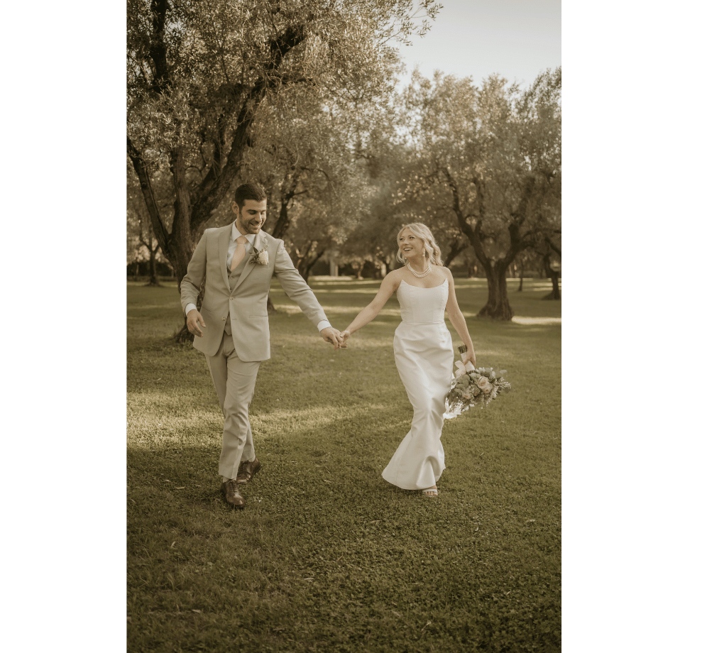 A bride and groom walk hand in hand on grass in a park, surrounded by trees, both smiling and dressed in formal wedding attire. This wedding was planned by Bweddings.