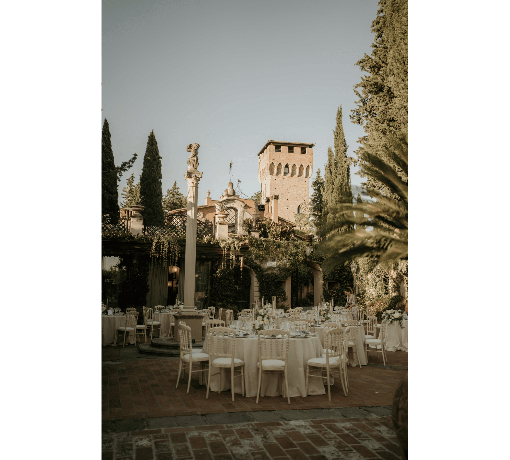 Outdoor courtyard set for an event with round dining tables and chairs, surrounded by greenery, columns, and a historic building in the background. This wedding was planned by Bweddings.