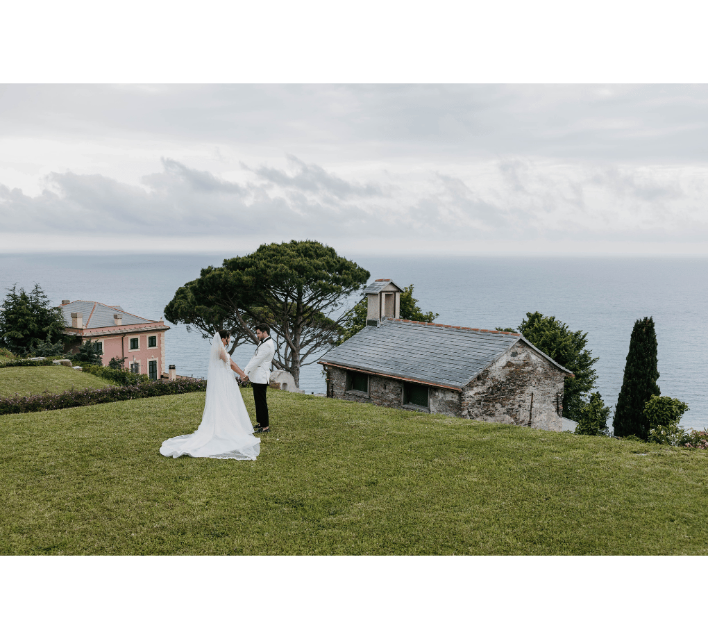 A bride and groom stand together on a grassy hill overlooking the sea, with a stone building and trees in the background.