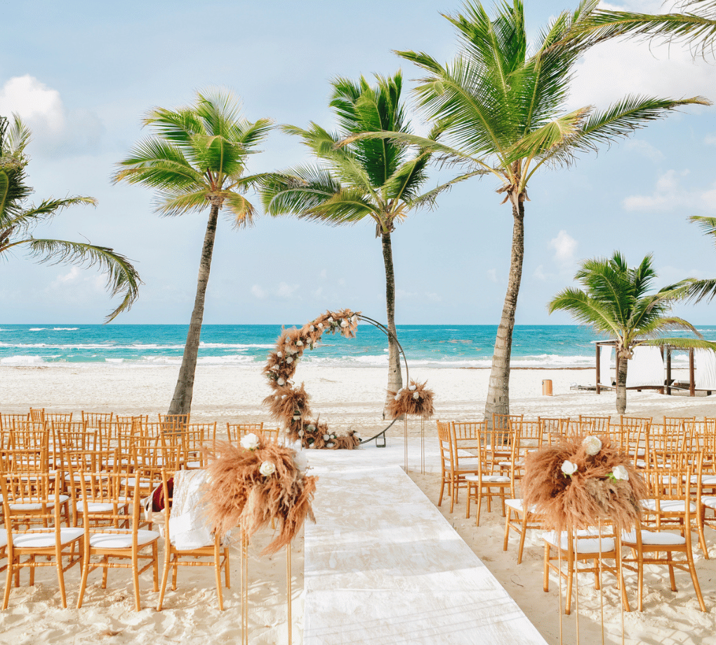 Beach wedding setup with wooden chairs facing an ocean backdrop, palm trees, and a decorated circular arch under a clear sky.