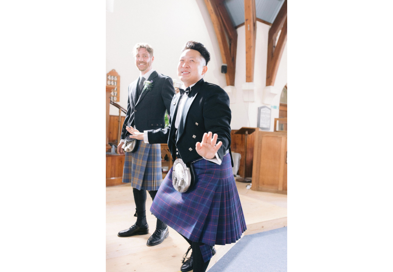 A groom and a wedding planner wearing kilts and formal jackets stand smiling inside a church with wooden beams and light streaming in.